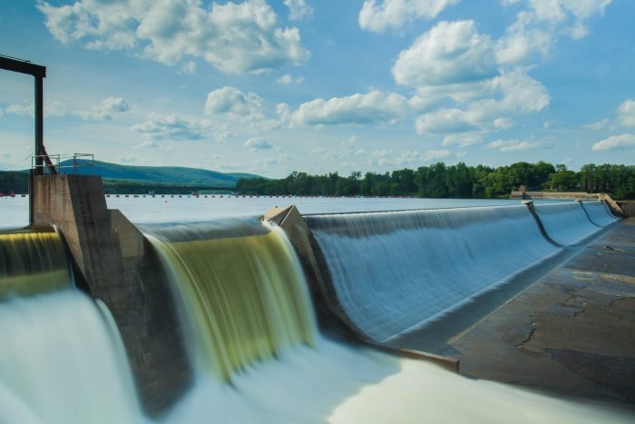 Uma vista aérea de uma barragem, com a água a fluir através das suas comportas e um vasto reservatório por trás.